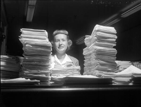 A woman smiles, sitting behind a desk between two tall stacks of paper 