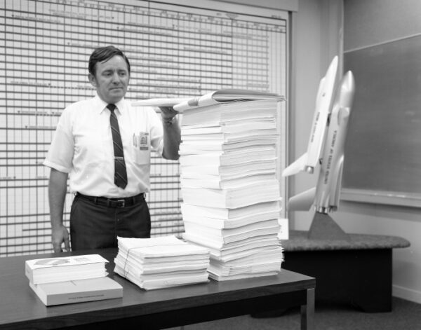 A man stands next to a pile of papers stacked on his desk, there is a model of a plane or space shuttle behind him