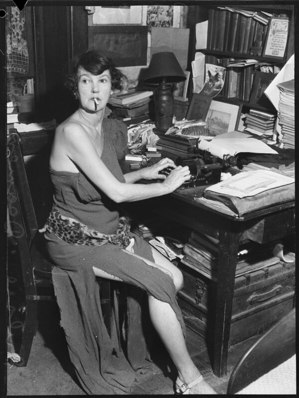 a woman in a fancy dress siting at a typewriter with a cigarette in her mouth. She is lookig over her shoulder ta the camera