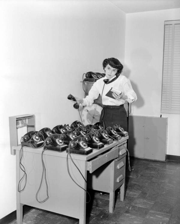 A woman wearing a white shirt and a dark skirt holds a phone to her ear while the desk next to her is full of ten other phones.