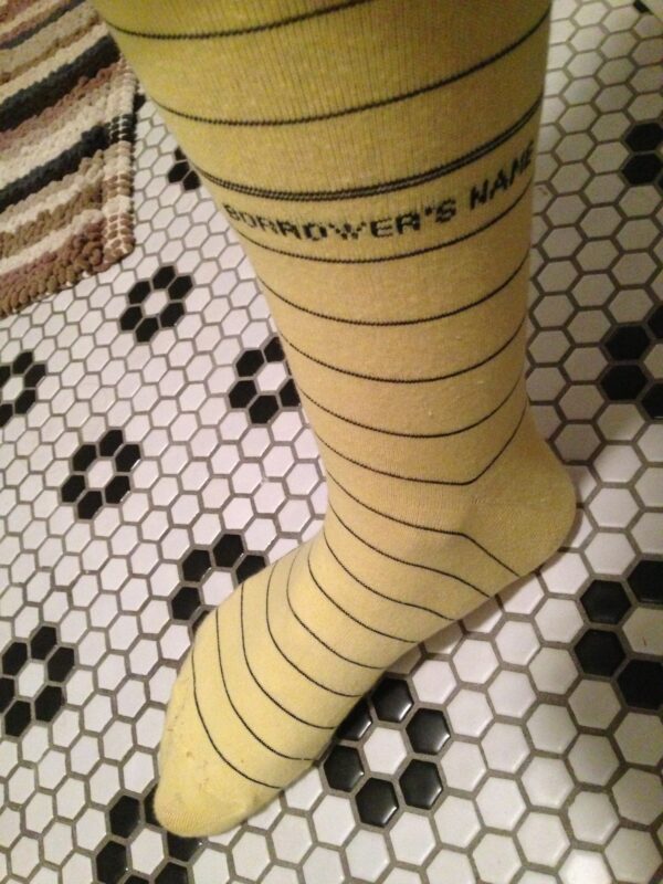 a foot in a sock that is yellow with thin black stripes and it meant to evoke a checkiout card in a book. It's standing on a tiled floor with black and white colored hexagons.